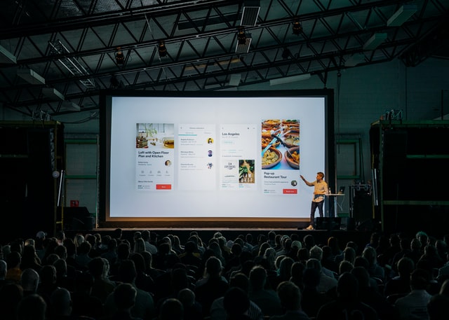 Man in yellow shirt points to large screen showing slides in front of large, dimmed audience