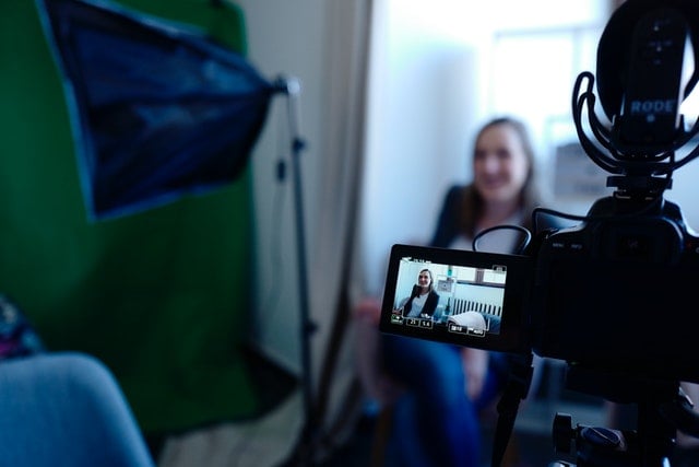 Woman sits behind a camera smiling on a set with green screen nearby